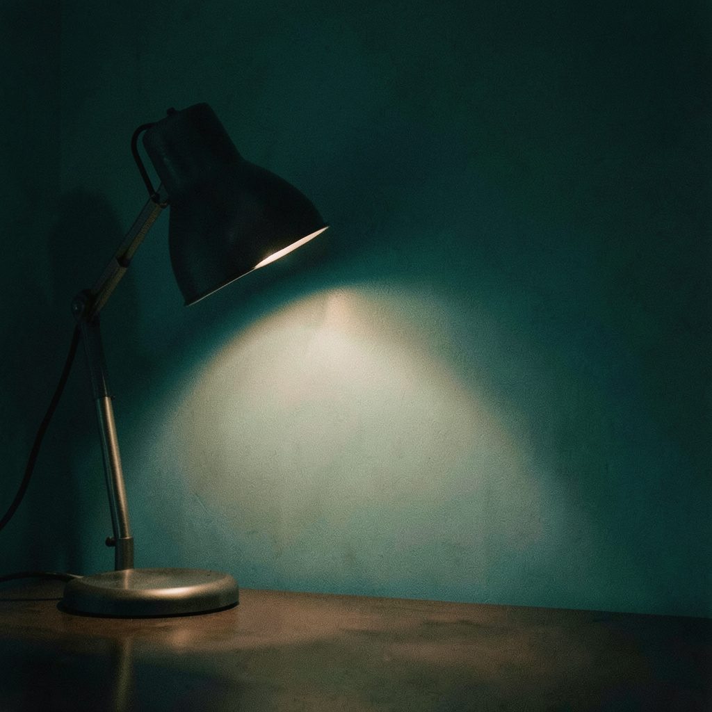 Duotone photograph of a dim room with a single desk lamp casting warm sepia light against a deep teal wall, moody and atmospheric.